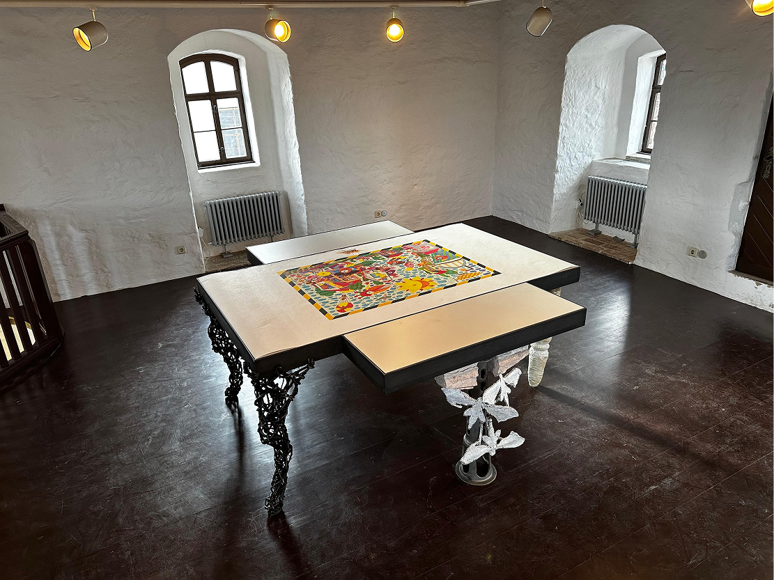 In einem historischen Raum mit dicken weißen Wänden und dunklem Boden steht ein Kunsttisch mit einer zentral platzierten, farbintensiven Illustration.