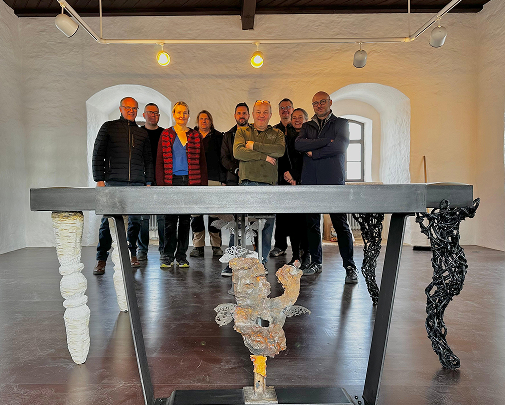 Zehn Menschen stehen in einem Raum mit weißen Wänden und Holzboden, im Vordergrund ein künstlerischer Tisch mit plastischen Elementen.